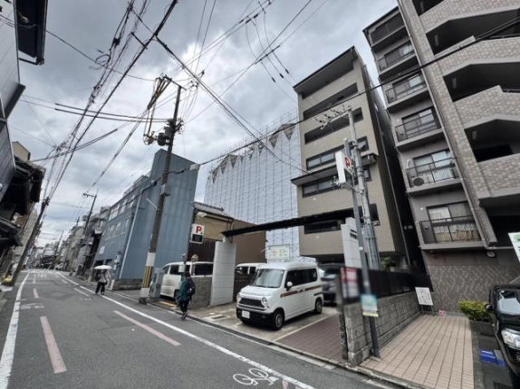 前面道路から見上げたビルと門まわり。左手には足場に覆われた建物と、自転車レーンのある通りが奥へ延びる