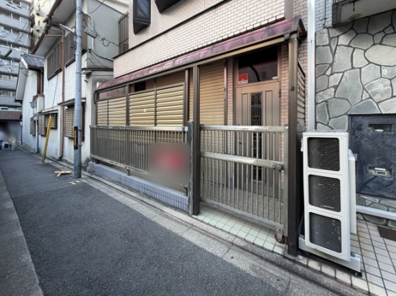 建物1階の入口まわり。ベージュのタイル外壁の下に格子の門扉とルーバーの目隠しがあり、奥に装飾ガラス入りの玄関ドア、右側に縦型の室外機がある