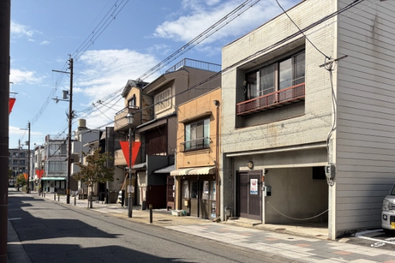 町家や商店が並ぶ北区の通り沿いに建つ貸店舗を斜めから見た様子。歩道には街灯と赤い三角フラッグが続く