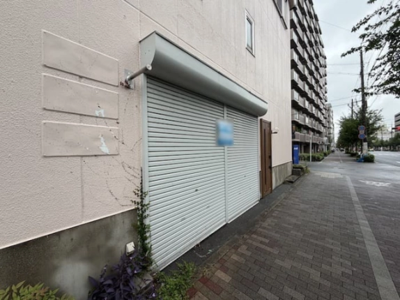 店舗正面を反対側から見た外観。淡い色の外壁とシャッター、右奥へ延びる歩道