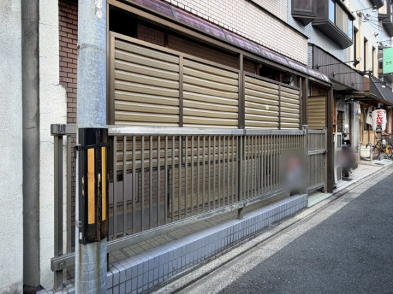 電柱越しに見た1階の格子フェンスとルーバー塀。タイル張りの腰壁沿いに路地が右奥へ続き、提灯や看板を掲げた店が並ぶ