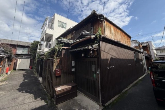 路地の入口付近。角に茶色の外壁と格子戸の木造2階建てが建ち、左手へ板塀沿いの路地が続く