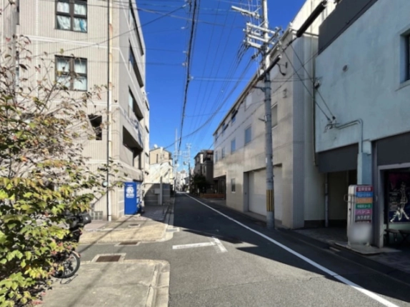 ビル前の通りの様子。右手に白いシャッターのある建物と電柱、左手に植え込みと青い自動販売機があり、道が奥へまっすぐ延びる