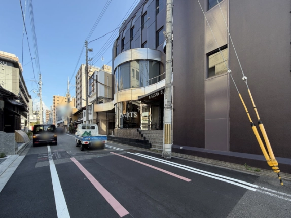 前面道路から見た建物と周辺の街並み。右側に建物が建ち、通りには車が停まり、奥に中層マンションが続く