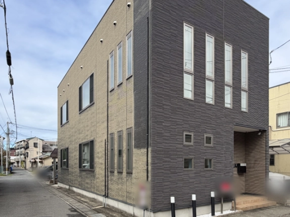 左斜め前から見た外観。ベージュの側面外壁に縦長窓と横長窓が並び、濃いグレーの正面右手に玄関、手前に黒い車止めポールが立つ