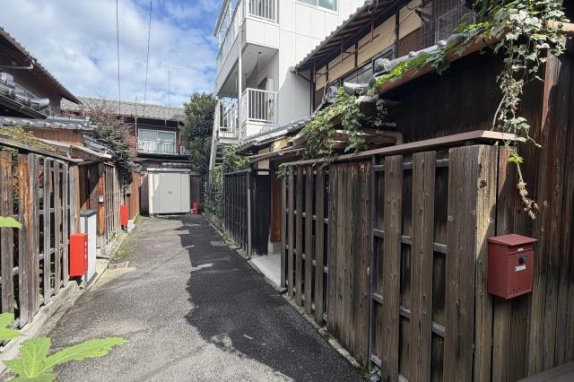 京町家の板塀沿いに奥へ延びる細い路地。蔦の絡む右側の塀に赤い郵便受けが付き、突き当たりに白い物置が見える