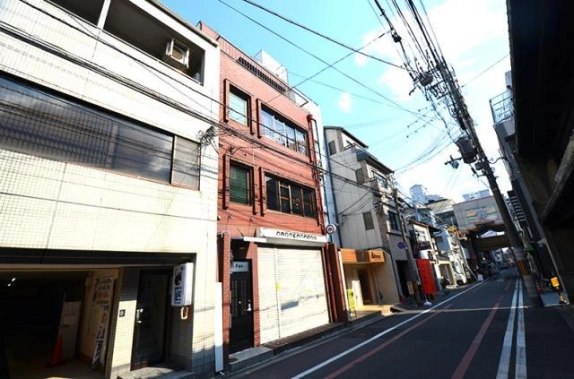 2階に202号室の店舗事務所がある建物を左斜め前から見た外観。白いビルの間に建ち、右へ延びる通りの先にアーケードが見える