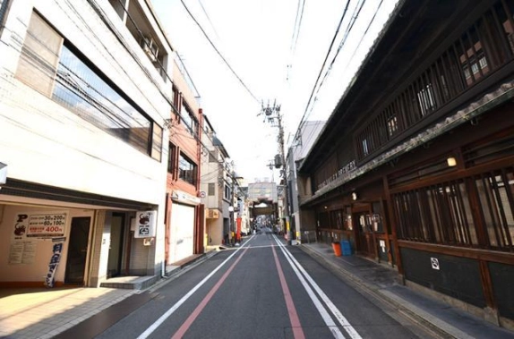 烏丸御池駅徒歩4分の中京区の前面道路を、反対方向に見通した様子。右に木格子の町家、左に駐車場のある白いビルが並び、奥にアーケードが見える