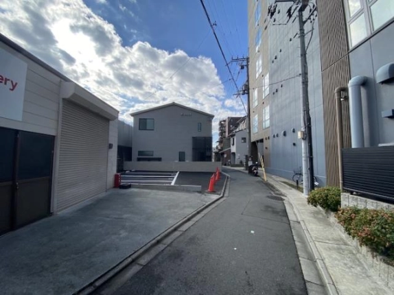 路地の先に白線を引いた舗装スペースとグレーの切妻屋根の2階建住宅が見える南区の街並み。左にシャッター付き倉庫、右に高い建物と電柱が続く