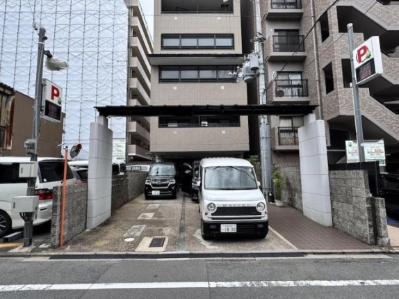 白い門柱と黒い梁の門構えを正面から見た様子。奥に1階区画前の敷地内駐車場が続き、右側はタイル張りの通路