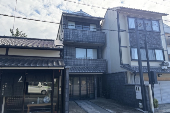 正面やや左から見た外観。暖簾の掛かった左隣の町家と右隣の3階建てに挟まれて、タイル張りの建物が建つ