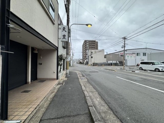 入口前から歩道沿いに見た前面道路。白線のある通りの先に中層マンション、道路の向かい側に駐車場が見える