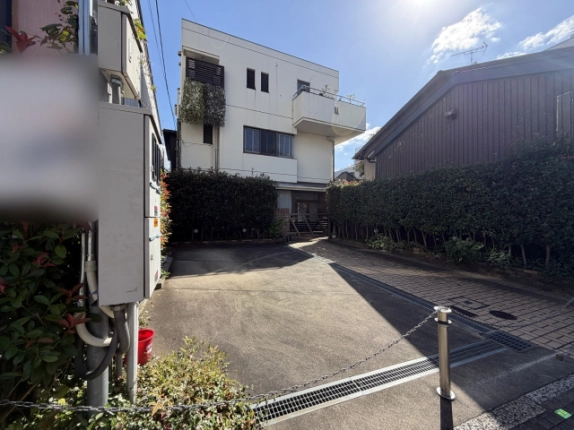 道路側から左斜めに見た建物と敷地。手前左に設備ボックス、チェーンを張ったポール、右奥に濃い茶色の板張りの建物