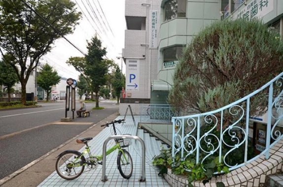 建物前のタイル張りの歩道。手前に駐輪ラックと自転車、右に水色の曲線状の装飾手すりと植栽、左に街路樹が並ぶ車道とバス停が見える