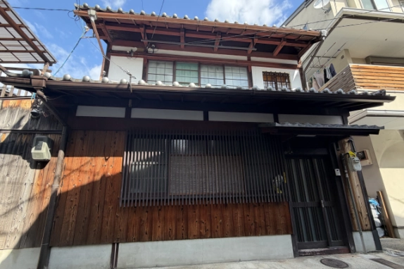 正面から見上げた町家の外観。2階は白壁にガラス窓と木格子の小窓、1階は板張りの壁に縦格子の窓が並び、右端に玄関がある