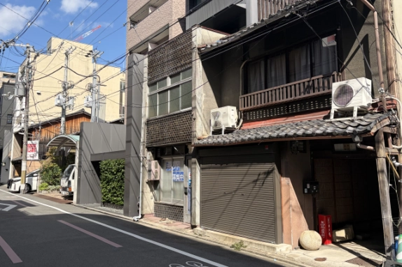 右斜め前から見た外観。手前に瓦の庇とシャッターがある隣の町家、物件の左に灰色の建物と植栽、奥に中京区の街並みが見える