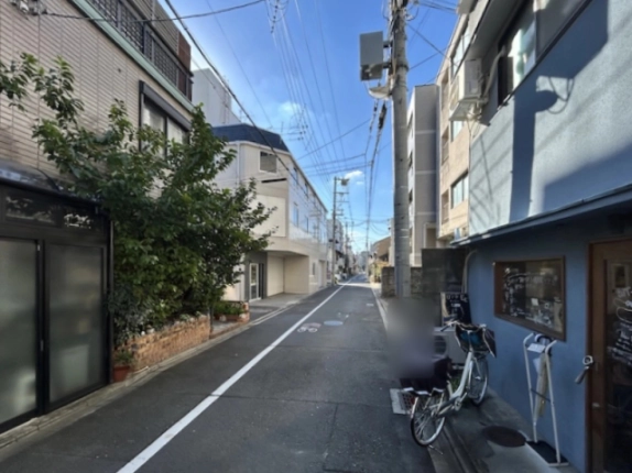 少し離れた通りから見たビルの位置。道路左手奥に紺色屋根の当ビル、手前左に植栽のある住宅、右手に青い外壁の建物と自転車