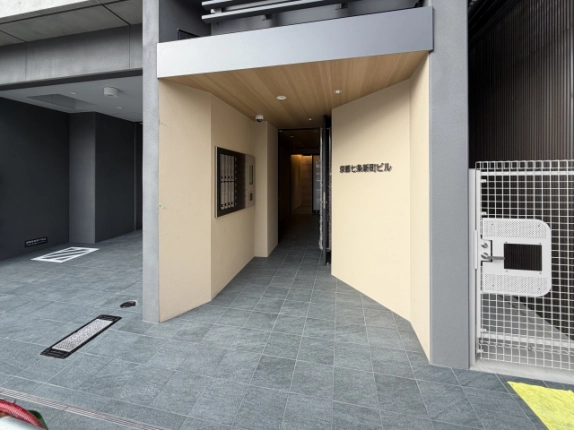 ベージュの壁と木目調の天井のエントランス。左の壁に集合ポスト、右の壁に館名サインがあり、奥へ通路が続く。床はグレーのタイル