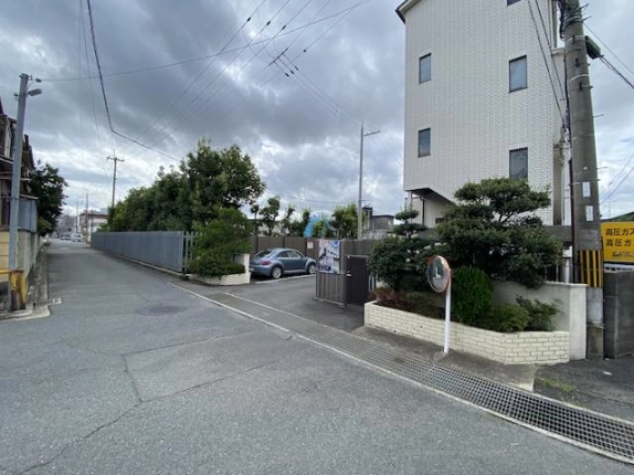 建物前の道路と出入口付近。右手に白い外壁の建物と植え込み、カーブミラーがあり、門扉の奥には車の停まる駐車スペースが見える