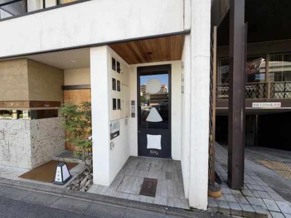丸・三角・四角のマークがあるビル入口。黒い扉と木目の天井、左手の植栽