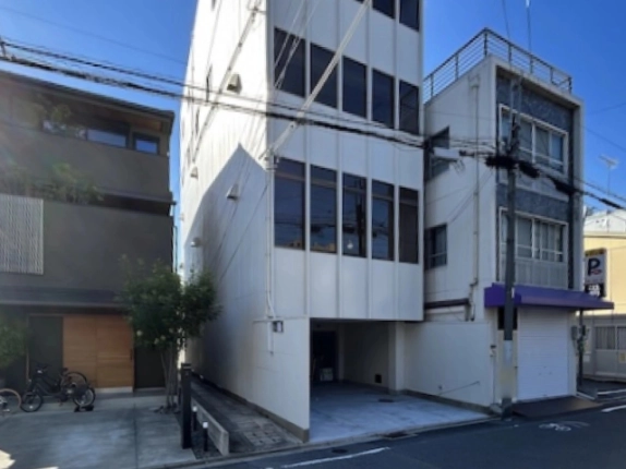 大宮通沿いに建つ白い外壁の5階建テナントビル。1階はピロティ状の駐車スペースで、左に黒い外壁の住宅、右に紫色の庇の建物が並ぶ