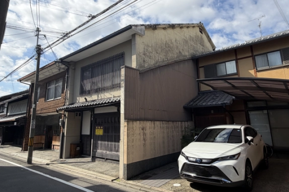 右斜め前から見た木造2階建の町家外観。右隣との間には波板張りの外壁が立ち、隣のカーポートに白い車が停まっている
