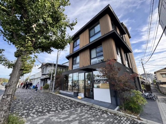 左京区田中下柳町、川端通に面した角地に建つ2021年築のビル外観。木目調の縦格子と黒い軒が重なる外壁を、イチョウ並木の歩道から見上げた様子