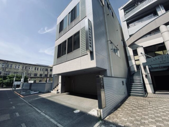 右斜め前から見た外観。壁面に縦長の袖看板、車庫の前に銘板が立ち、左奥には道路沿いの駐車場と古い洋風建築が見える