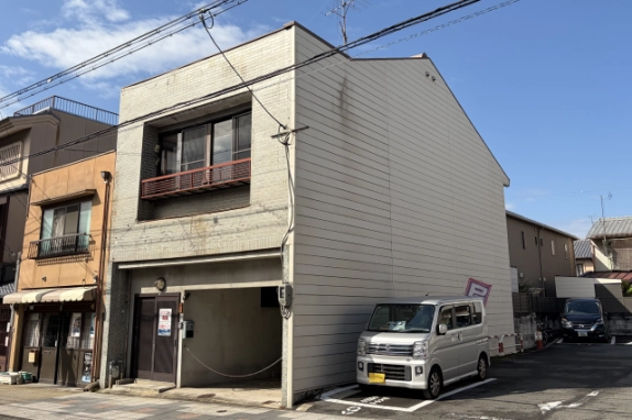 右斜め前から見た外観。ベージュの横張りの側面外壁と、隣の駐車場に停まるシルバーの軽バン