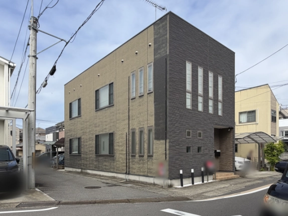 川島滑樋町一棟を交差点側から見た2階建ての箱型外観。ベージュと濃いグレーの2色の外壁で、左手前に電柱、建物の前に道路が広がる