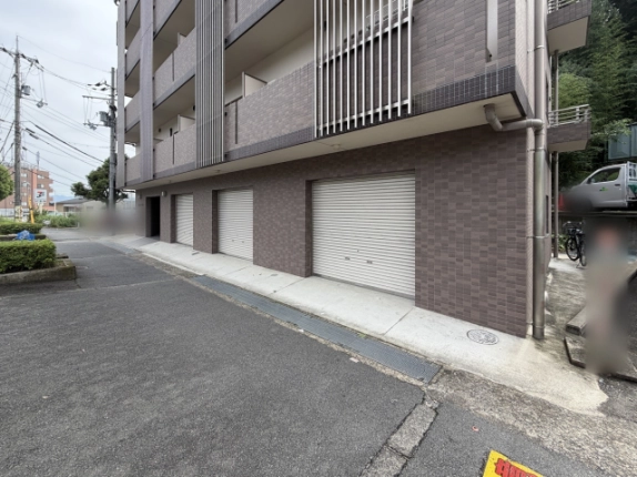 建物の角から斜めに見た1階部分。幅の異なる3つのシャッターが道路沿いに並び、手前の歩道際に側溝のグレーチングが続く