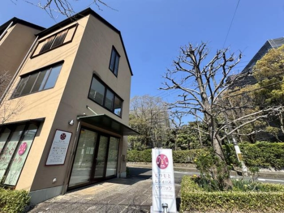 伊佐ビルの角を反対側から見た外観。二面の窓と緑のひさし、右手の道路沿いの樹木