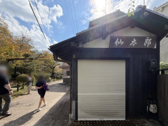 妻側の1階シャッターと上部の木製看板、黒い板張りの外壁。左手に人が行き交う砂利道