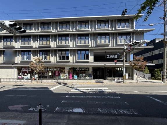左京区の2waを正面から見た外観。打放しコンクリートの4階建てで、1階にスーパーマーケット、右寄りに「2wa」の看板を掲げた通路状の入口、手前に横断歩道