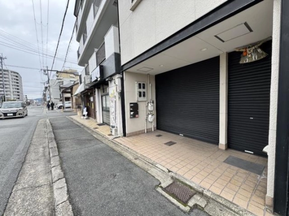 左側から見た1階入口まわり。黒いシャッター2枚とタイル張りの床があり、歩道沿いの左手に隣の店舗が並ぶ