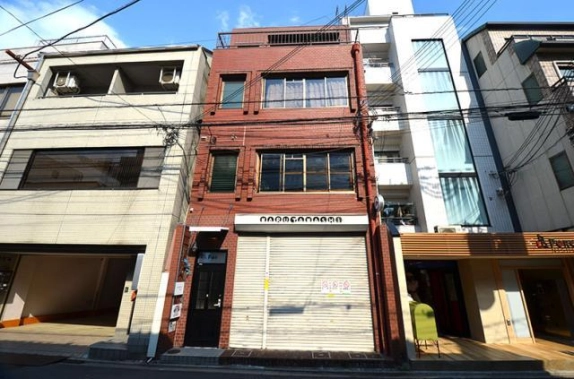 正面から見た赤レンガ調の外壁。1階はシャッターと、その左手にあるガラス入りの黒いドアの入口。2階と3階には木枠の窓が並ぶ