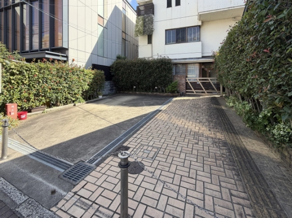 道路際から見た敷地の入口。ヘリンボーン状のレンガ敷き通路と排水溝の格子、左手に消火器箱と赤いバケツ