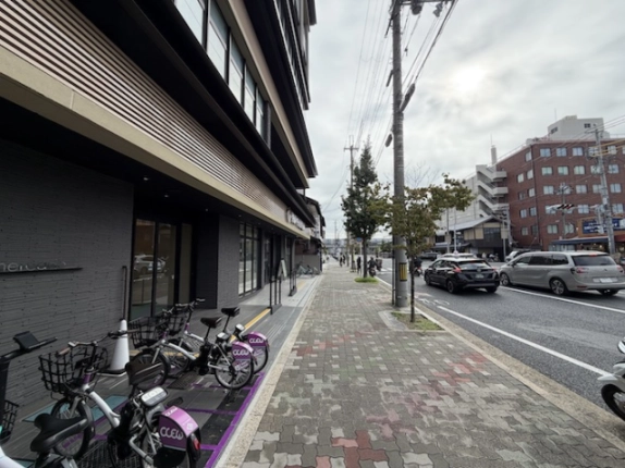 建物を左手に見た前面道路。店舗前に並ぶ自転車と街路樹のある歩道