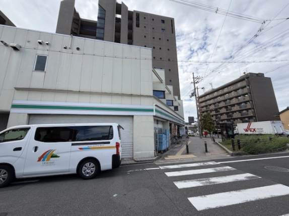 建物の角と側道を見た様子。白い外壁の手前に車が停まり、右手に横断歩道がある