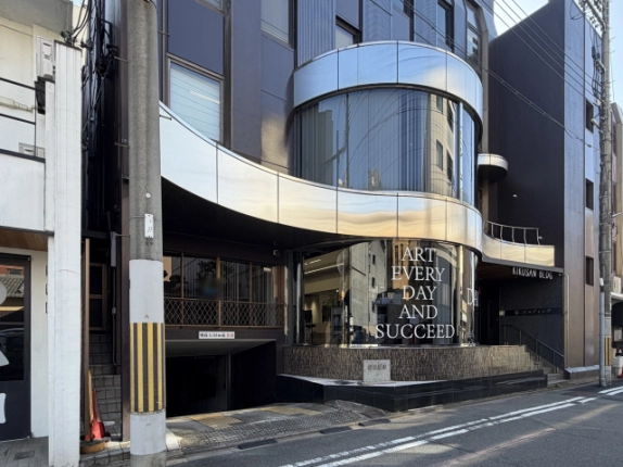 左斜め前から見た菊三ビル。円筒形のガラス窓と曲線の庇の下に石の銘板があり、左手に車高制限表示のある地下駐車場の入口が見える