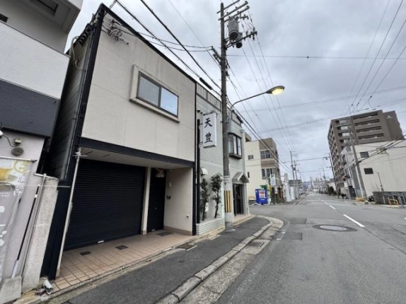 2階に窓がある2階建てのベージュの建物。1階に黒いシャッターがあり、右手には街灯の立つ道路が奥へ延びる
