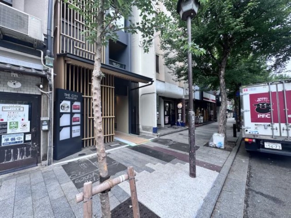 木屋町通の歩道から斜めに見たTHE CITY 先斗町の外観。木製の縦格子と黒い館名サイン、奥へ延びる入口通路、右手に街路樹が並ぶ