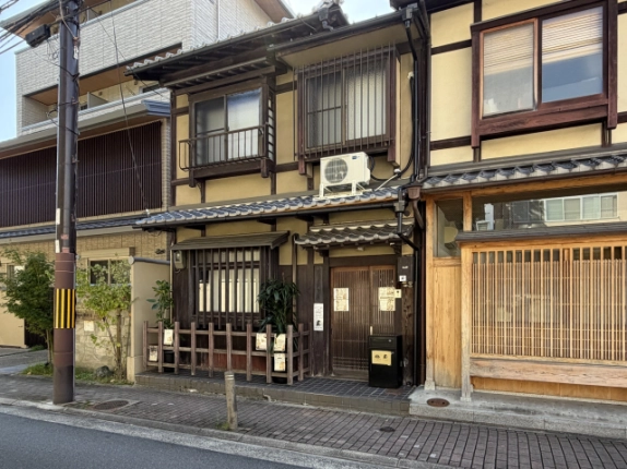 左斜め前から見た2階建て町家の外観。2階に手すり付きの窓と縦格子の窓が並び、1階には瓦屋根の庇の下に格子窓と玄関がある