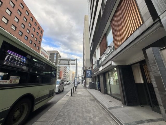 建物を右手に見た四条通の歩道。縦格子のある低層部と車道を走るバス