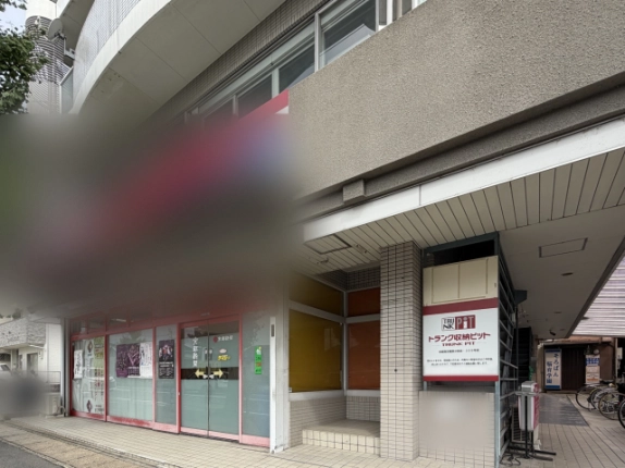 渡邉ビルの1階正面。左に赤い枠のガラス張り店舗、右に黄色とオレンジのブラインド窓とタイル張りの段差がある奥まった入口