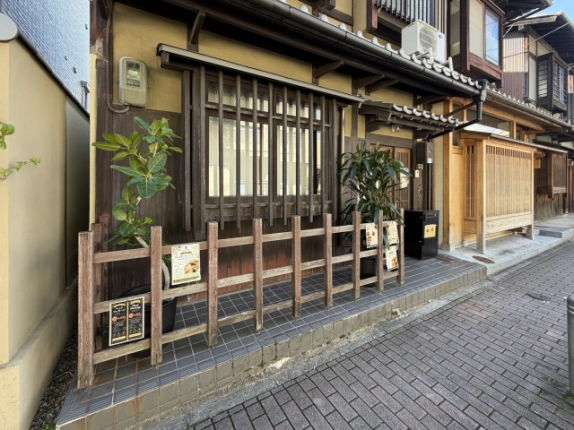 1階正面左側の縦格子の窓と木製の柵。柵の内側に鉢植えの観葉植物があり、足元はタイル張りで歩道に面している