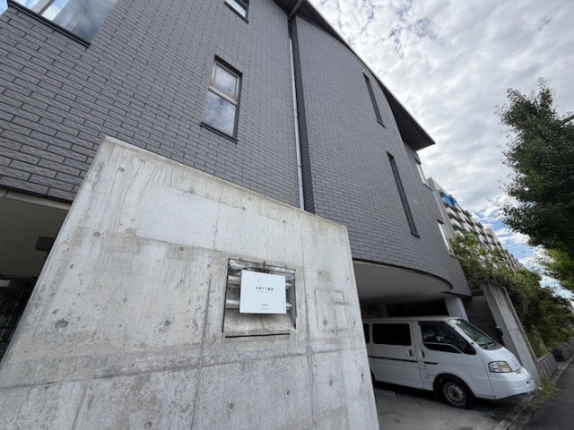 建物を反対側から見上げた様子。手前のコンクリート壁と灰色の外壁、1階の駐車スペース