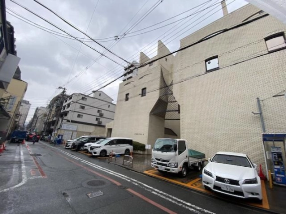 左手に通りの奥まで見通せる位置から見たビルの外観。右側に白いタイル壁の建物があり、道路沿いの駐車区画にトラックや乗用車が並ぶ