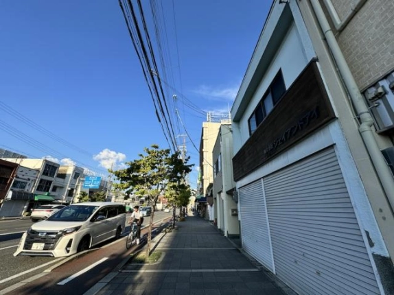 建物前の歩道から七条通を見通した広角の眺め。右端に看板帯とシャッター、左の車道に車と自転車、青空に電線が延びる