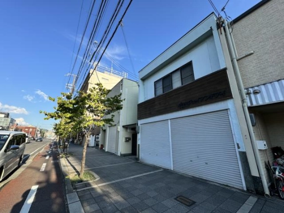 梅小路京都西駅徒歩6分、七条通の歩道から斜めに見た建物。右に白い外壁とシャッター、左に街路樹が並ぶ広い歩道と自転車レーンのある車道